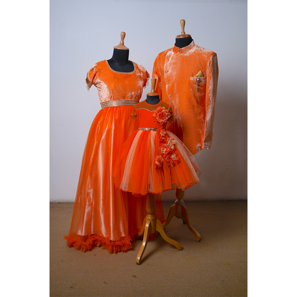 Family matching outfit set in vibrant orange velvet & net - includes mother’s gown, father’s sherwani jacket, and girl’s frock with floral appliqué.