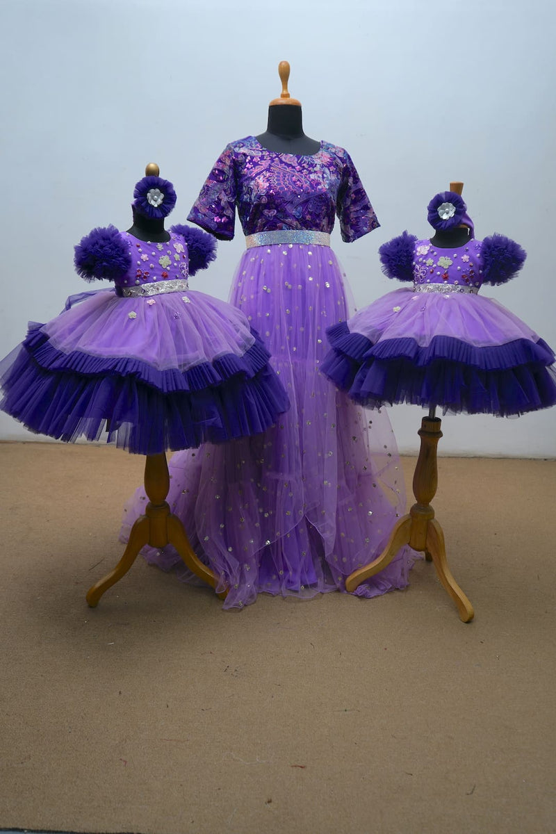 Regal purple matching family gown set featuring shimmer net Mom's gown and two baby girl layered gowns with floral appliqué and headbands.