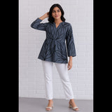 Woman wearing a charcoal grey flared top with black leaf prints, paired with white pants, posing against a white brick wall.