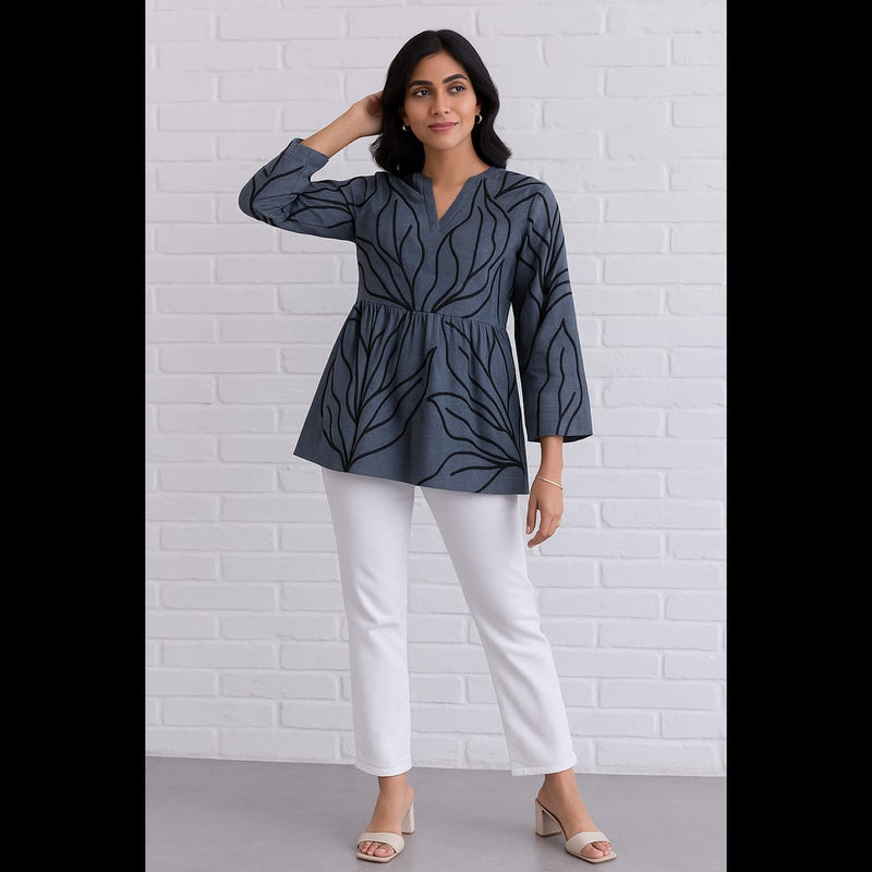 Woman wearing a charcoal grey flared top with black leaf prints, paired with white pants, posing against a white brick wall.