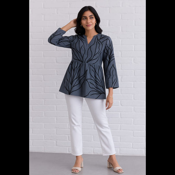 Woman wearing a charcoal grey flared top with black leaf prints, paired with white pants, posing against a white brick wall.