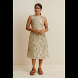 Woman wearing a sleeveless beige dress with black leaf prints, standing indoors in a minimal setup.